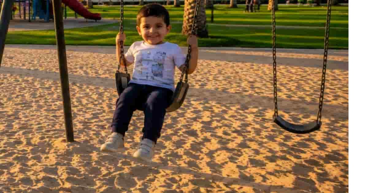 park with a colorful playground, palm trees, green lawns, walking paths, and a child on a swing.